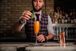 A bartender dressed in a plaid vest and a tie is skillfully preparing a cocktail at a bar. He is carefully placing a mint leaf garnish on top of an orange-colored drink served in a champagne flute. The background displays an array of liquor bottles on shelves and copper cocktail shakers on the counter.