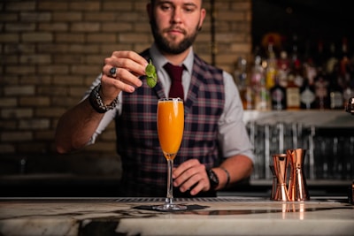 A bartender dressed in a plaid vest and a tie is skillfully preparing a cocktail at a bar. He is carefully placing a mint leaf garnish on top of an orange-colored drink served in a champagne flute. The background displays an array of liquor bottles on shelves and copper cocktail shakers on the counter.