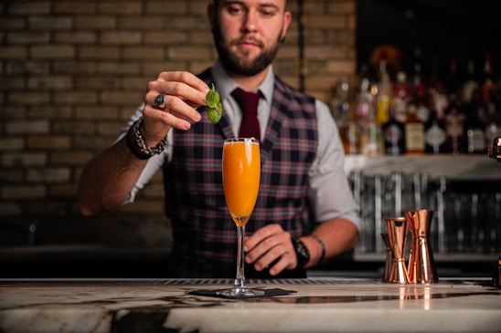 A bartender dressed in a plaid vest and a tie is skillfully preparing a cocktail at a bar. He is carefully placing a mint leaf garnish on top of an orange-colored drink served in a champagne flute. The background displays an array of liquor bottles on shelves and copper cocktail shakers on the counter.