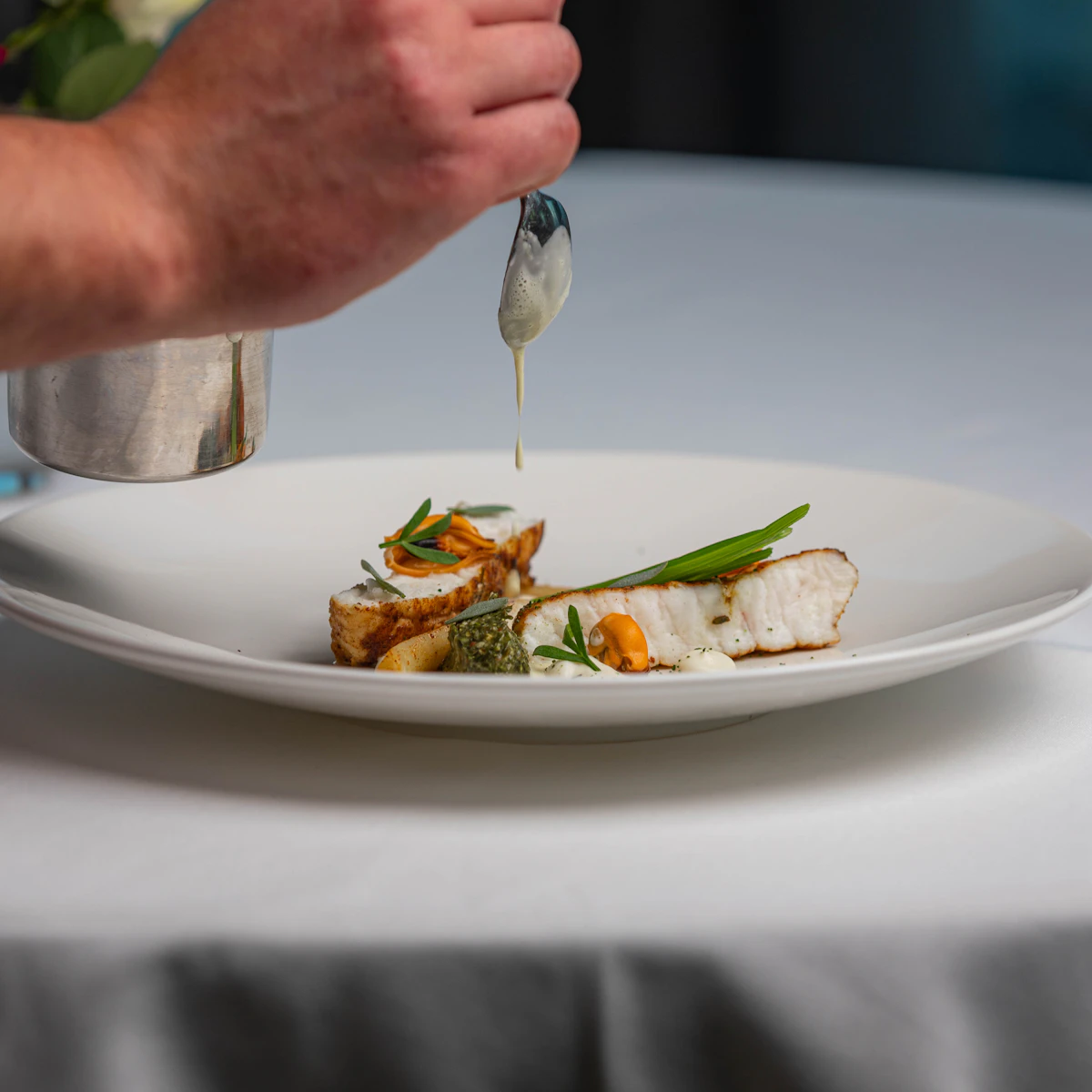 Uno chef versa una salsa su un filetto di pesce impiattato con verdure, su tovaglia bianca