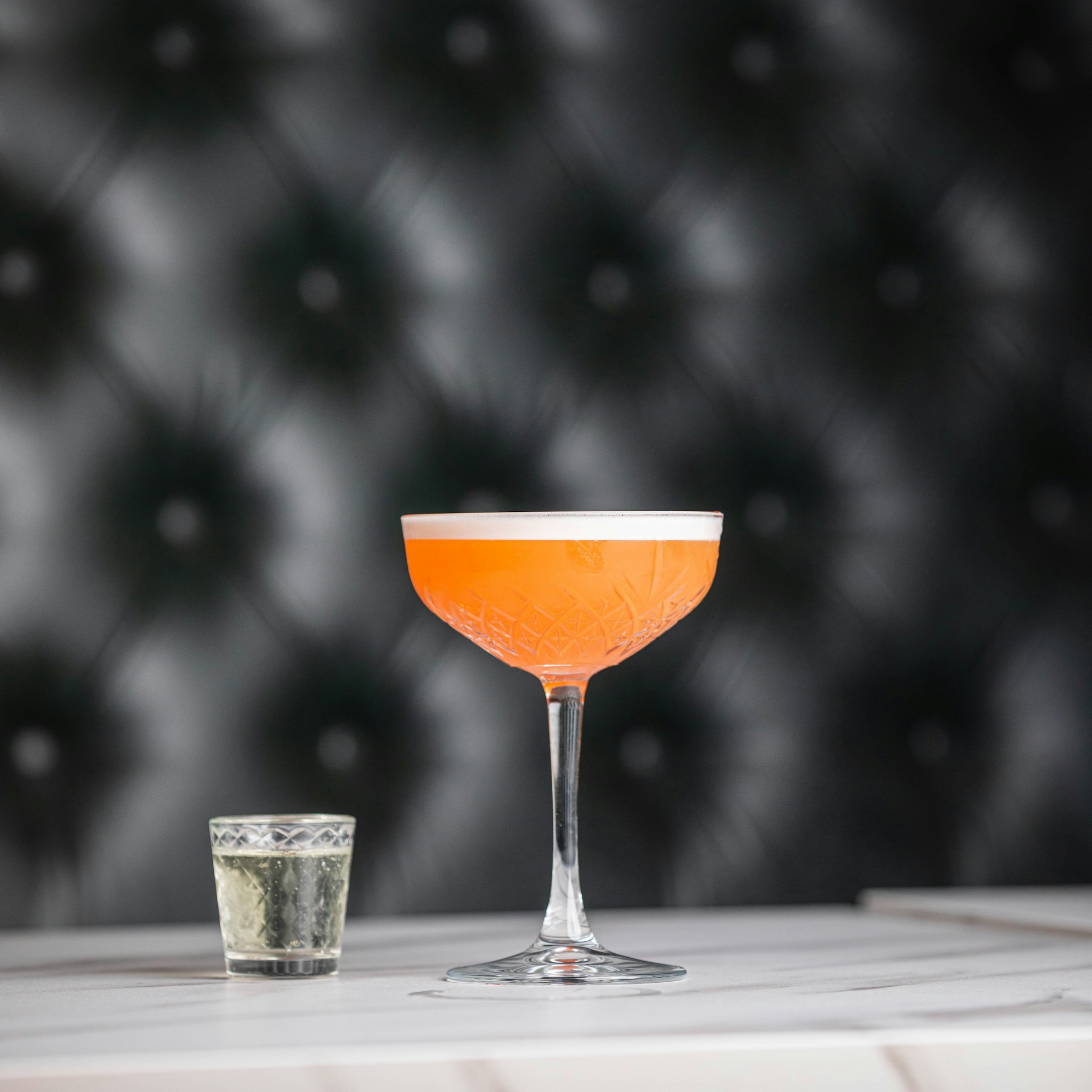 An elegant cocktail in a crystal glass, garnished with a twist of orange peel, glowing softly against a backdrop of black and cream tones with delicate Mexican tile motifs.