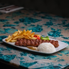A white rectangular plate is arranged with seasoned grilled meat skewers, a serving of white rice, crispy golden french fries, and slices of red and green bell peppers. The plate is set on a colorful marble table with blue and beige patterns.