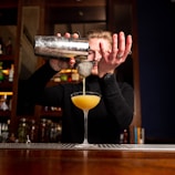 A bartender is skillfully pouring a yellow cocktail from a shaker through a strainer into a cocktail glass. The background features shelves lined with bottles, suggesting a well-stocked bar setting. The focus is on the bartender's hands and the pouring action, capturing a moment of precision in mixology.