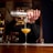 A bartender is skillfully pouring a yellow cocktail from a shaker through a strainer into a cocktail glass. The background features shelves lined with bottles, suggesting a well-stocked bar setting. The focus is on the bartender's hands and the pouring action, capturing a moment of precision in mixology.