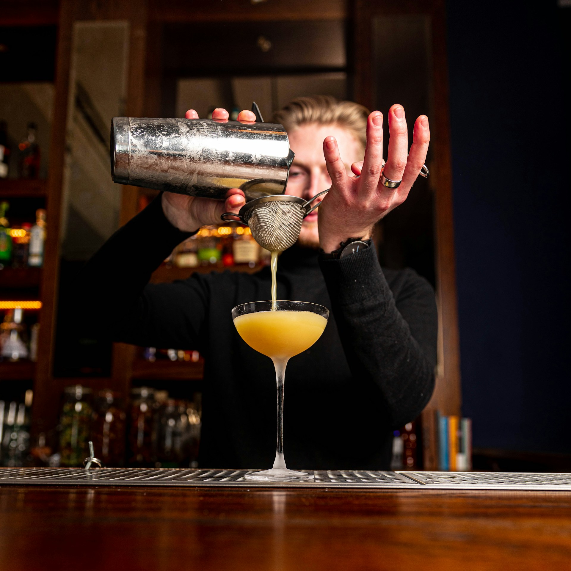 A vibrant bartender skillfully pouring a colorful cocktail into a martini glass at a lively event.