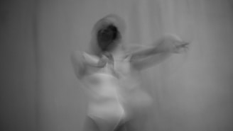 A black and white image depicting a blurred figure in motion, conveying a sense of fluidity and movement. The figure appears to be dancing or in an expressive pose, with an indistinct form creating a ghostly or ethereal effect.