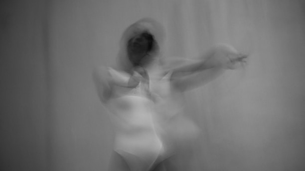A black and white image depicting a blurred figure in motion, conveying a sense of fluidity and movement. The figure appears to be dancing or in an expressive pose, with an indistinct form creating a ghostly or ethereal effect.