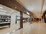 A modern store with a sleek interior is located inside a shopping mall. The store features shelves displaying various products like phone cases and accessories. There is a central table with seating, and a reception desk nearby. The mall corridor appears expansive with a polished floor and muted lighting, giving a luxurious feel. Several people are walking in the distance along the corridor.