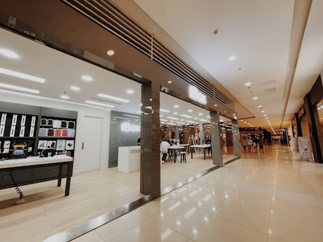 A modern store with a sleek interior is located inside a shopping mall. The store features shelves displaying various products like phone cases and accessories. There is a central table with seating, and a reception desk nearby. The mall corridor appears expansive with a polished floor and muted lighting, giving a luxurious feel. Several people are walking in the distance along the corridor.