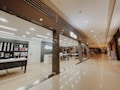 A modern store with a sleek interior is located inside a shopping mall. The store features shelves displaying various products like phone cases and accessories. There is a central table with seating, and a reception desk nearby. The mall corridor appears expansive with a polished floor and muted lighting, giving a luxurious feel. Several people are walking in the distance along the corridor.