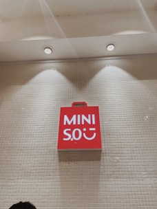 A red sign with white text reading 'MINISO' is mounted on a tiled wall under a ceiling with recessed lights.