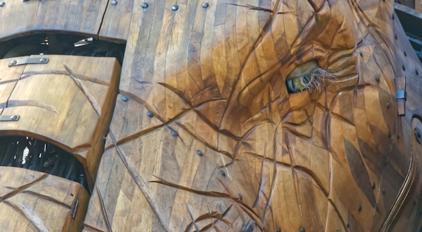 A close-up of an intricately detailed wooden structure resembling the head of an animal, possibly an elephant. The craftsmanship displays various wooden panels with visible bolts and joins, giving it an articulated and lifelike appearance. The eye, made from synthetic materials, adds a realistic touch.