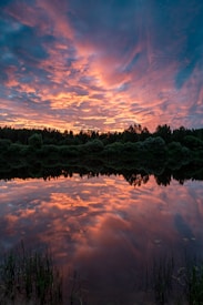 A serene landscape featuring a vibrant sunset with dramatic clouds that reflect beautifully on a still lake. The sky is filled with shades of orange, pink, and purple, creating a stunning contrast against the silhouette of dense trees lining the horizon.