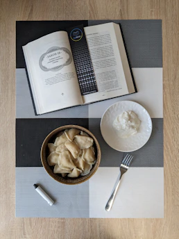 An open book with a bookmark is placed on a checkered table mat. Beside it, a bowl filled with dumplings is accompanied by a plate with a white sauce and a fork. A small cylindrical object, possibly a lighter, is also on the table. The background is a wooden surface.