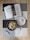An open book with a bookmark is placed on a checkered table mat. Beside it, a bowl filled with dumplings is accompanied by a plate with a white sauce and a fork. A small cylindrical object, possibly a lighter, is also on the table. The background is a wooden surface.