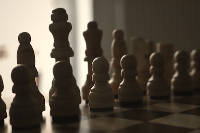Silhouettes of chess pieces are arranged on a board, illuminated from behind. The pieces create a dramatic contrast between light and dark, with a minimalistic, shadowy appearance that emphasizes their distinctive shapes.