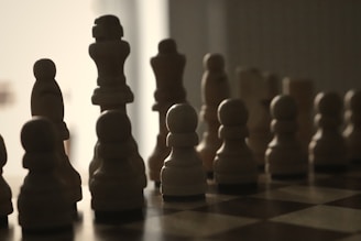 Silhouettes of chess pieces are arranged on a board, illuminated from behind. The pieces create a dramatic contrast between light and dark, with a minimalistic, shadowy appearance that emphasizes their distinctive shapes.