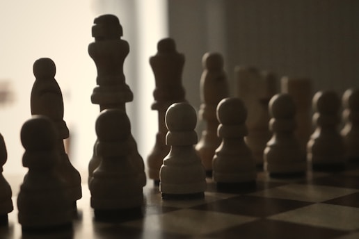 Silhouettes of chess pieces are arranged on a board, illuminated from behind. The pieces create a dramatic contrast between light and dark, with a minimalistic, shadowy appearance that emphasizes their distinctive shapes.