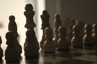 Silhouettes of chess pieces are arranged on a board, illuminated from behind. The pieces create a dramatic contrast between light and dark, with a minimalistic, shadowy appearance that emphasizes their distinctive shapes.