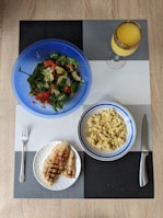 A meal is arranged on a textured placemat with geometric patterns. A vibrant blue bowl contains a fresh salad with leafy greens, diced tomatoes, cucumbers, and avocado. Next to it is a bowl filled with cooked grains, likely quinoa or bulgur, in a white and blue-rimmed bowl. A plate features grilled chicken breast with grill marks, placed beside a fork. A decorative glass holds an orange beverage, possibly juice.