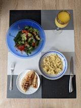 A meal is arranged on a textured placemat with geometric patterns. A vibrant blue bowl contains a fresh salad with leafy greens, diced tomatoes, cucumbers, and avocado. Next to it is a bowl filled with cooked grains, likely quinoa or bulgur, in a white and blue-rimmed bowl. A plate features grilled chicken breast with grill marks, placed beside a fork. A decorative glass holds an orange beverage, possibly juice.
