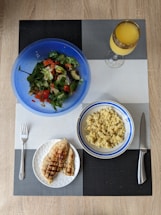 A meal is arranged on a textured placemat with geometric patterns. A vibrant blue bowl contains a fresh salad with leafy greens, diced tomatoes, cucumbers, and avocado. Next to it is a bowl filled with cooked grains, likely quinoa or bulgur, in a white and blue-rimmed bowl. A plate features grilled chicken breast with grill marks, placed beside a fork. A decorative glass holds an orange beverage, possibly juice.