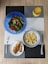 A meal is arranged on a textured placemat with geometric patterns. A vibrant blue bowl contains a fresh salad with leafy greens, diced tomatoes, cucumbers, and avocado. Next to it is a bowl filled with cooked grains, likely quinoa or bulgur, in a white and blue-rimmed bowl. A plate features grilled chicken breast with grill marks, placed beside a fork. A decorative glass holds an orange beverage, possibly juice.