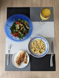 A meal is arranged on a textured placemat with geometric patterns. A vibrant blue bowl contains a fresh salad with leafy greens, diced tomatoes, cucumbers, and avocado. Next to it is a bowl filled with cooked grains, likely quinoa or bulgur, in a white and blue-rimmed bowl. A plate features grilled chicken breast with grill marks, placed beside a fork. A decorative glass holds an orange beverage, possibly juice.