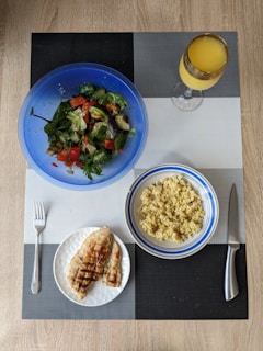 A meal is arranged on a textured placemat with geometric patterns. A vibrant blue bowl contains a fresh salad with leafy greens, diced tomatoes, cucumbers, and avocado. Next to it is a bowl filled with cooked grains, likely quinoa or bulgur, in a white and blue-rimmed bowl. A plate features grilled chicken breast with grill marks, placed beside a fork. A decorative glass holds an orange beverage, possibly juice.
