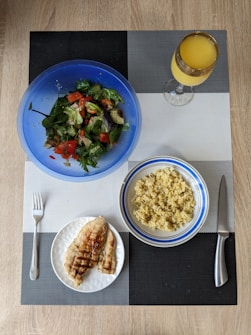 A meal is arranged on a textured placemat with geometric patterns. A vibrant blue bowl contains a fresh salad with leafy greens, diced tomatoes, cucumbers, and avocado. Next to it is a bowl filled with cooked grains, likely quinoa or bulgur, in a white and blue-rimmed bowl. A plate features grilled chicken breast with grill marks, placed beside a fork. A decorative glass holds an orange beverage, possibly juice.