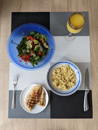 A meal is arranged on a textured placemat with geometric patterns. A vibrant blue bowl contains a fresh salad with leafy greens, diced tomatoes, cucumbers, and avocado. Next to it is a bowl filled with cooked grains, likely quinoa or bulgur, in a white and blue-rimmed bowl. A plate features grilled chicken breast with grill marks, placed beside a fork. A decorative glass holds an orange beverage, possibly juice.
