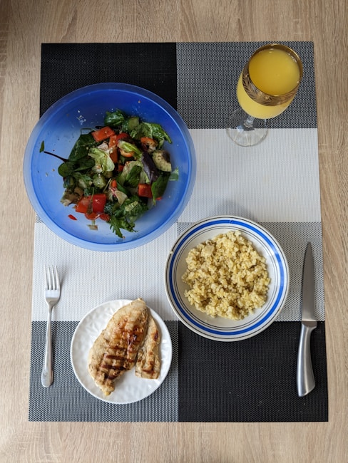 A meal is arranged on a textured placemat with geometric patterns. A vibrant blue bowl contains a fresh salad with leafy greens, diced tomatoes, cucumbers, and avocado. Next to it is a bowl filled with cooked grains, likely quinoa or bulgur, in a white and blue-rimmed bowl. A plate features grilled chicken breast with grill marks, placed beside a fork. A decorative glass holds an orange beverage, possibly juice.