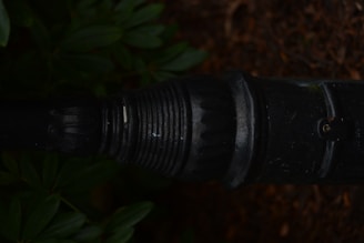 A close-up view of a dark, weathered metal pipe or connector with ribbed and indented textures. Surrounding the pipe are green leaves and brown soil, suggesting an outdoor setting.