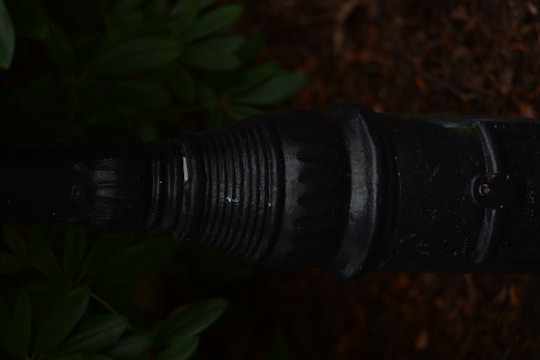 A close-up view of a dark, weathered metal pipe or connector with ribbed and indented textures. Surrounding the pipe are green leaves and brown soil, suggesting an outdoor setting.