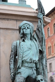 A verdigris-covered statue of a historical figure, likely a military leader or revolutionary, holding a large flagpole in one hand and a long staff or weapon in the other. The figure is dressed in detailed period attire, including a hat and a coat with buttons. The backdrop includes part of an ornate building with arched windows and decorative architectural elements.