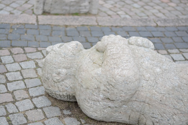 A stone sculpture depicting a reclining figure with hands folded on its chest, positioned on a cobblestone pavement. The texture of the stone is rough and pale, contrasting with the variety of dark and light gray tones of the cobblestones.