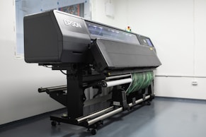 A large industrial printer is positioned in a clean, white-walled room. The printer is branded with the name Epson and appears to be printing or set up for a print job, with a green patterned material partially rolled through it. The room has a minimalistic look, with visible floor and wall fixtures.