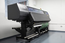 A large industrial printer is positioned in a clean, white-walled room. The printer is branded with the name Epson and appears to be printing or set up for a print job, with a green patterned material partially rolled through it. The room has a minimalistic look, with visible floor and wall fixtures.