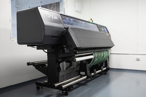A large industrial printer is positioned in a clean, white-walled room. The printer is branded with the name Epson and appears to be printing or set up for a print job, with a green patterned material partially rolled through it. The room has a minimalistic look, with visible floor and wall fixtures.