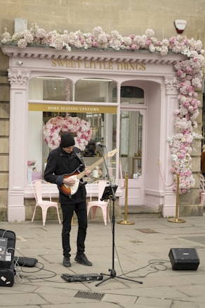 A person dressed in dark clothing plays an electric guitar in front of a storefront. The shop has pink and white floral decorations surrounding its entrance and a sign reading 'Sweet Little Things'. Several pink chairs are visible just inside the store's window display.