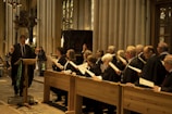 A group of people holding papers are dressed in formal robes and are standing in wooden pews inside a grand cathedral with high ceilings and stained glass windows. A man stands at a podium reading from a piece of paper.