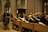 A group of people holding papers are dressed in formal robes and are standing in wooden pews inside a grand cathedral with high ceilings and stained glass windows. A man stands at a podium reading from a piece of paper.