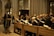 A group of people holding papers are dressed in formal robes and are standing in wooden pews inside a grand cathedral with high ceilings and stained glass windows. A man stands at a podium reading from a piece of paper.
