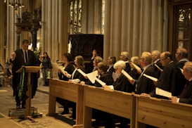 A group of people holding papers are dressed in formal robes and are standing in wooden pews inside a grand cathedral with high ceilings and stained glass windows. A man stands at a podium reading from a piece of paper.