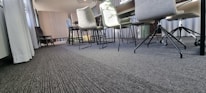 A modern conference room with a long table and multiple chairs on a carpeted floor. The chairs have different designs, including some with wheels. Sunlight streams through large windows covered by light curtains, and wall paneling adds texture to the background.