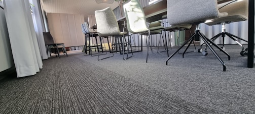 A modern conference room with a long table and multiple chairs on a carpeted floor. The chairs have different designs, including some with wheels. Sunlight streams through large windows covered by light curtains, and wall paneling adds texture to the background.