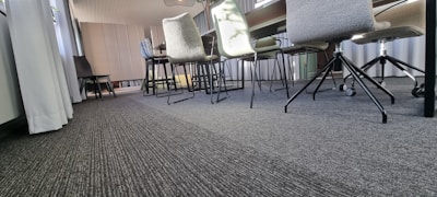 A modern conference room with a long table and multiple chairs on a carpeted floor. The chairs have different designs, including some with wheels. Sunlight streams through large windows covered by light curtains, and wall paneling adds texture to the background.