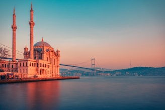 A mosque with two tall minarets is situated by a body of water. In the background, a large suspension bridge stretches across the scene with a hilly landscape in the distance. The sky appears during sunset or sunrise, with a gradient of warm colors.