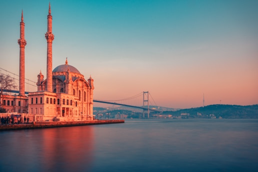 A mosque with two tall minarets is situated by a body of water. In the background, a large suspension bridge stretches across the scene with a hilly landscape in the distance. The sky appears during sunset or sunrise, with a gradient of warm colors.