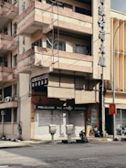 A multi-story building with a sun-worn façade and balconies rising above a closed clinic named 'Poliklinik Tan Lee & Cheong'. The building's exterior displays large Chinese characters, and wires run along the walls. Below, two motorcycles are parked beside the entrance, and a few utility boxes are positioned near the street corner. A traffic sign stands nearby, indicating a sharp turn. The environment appears urban with minimal activity.